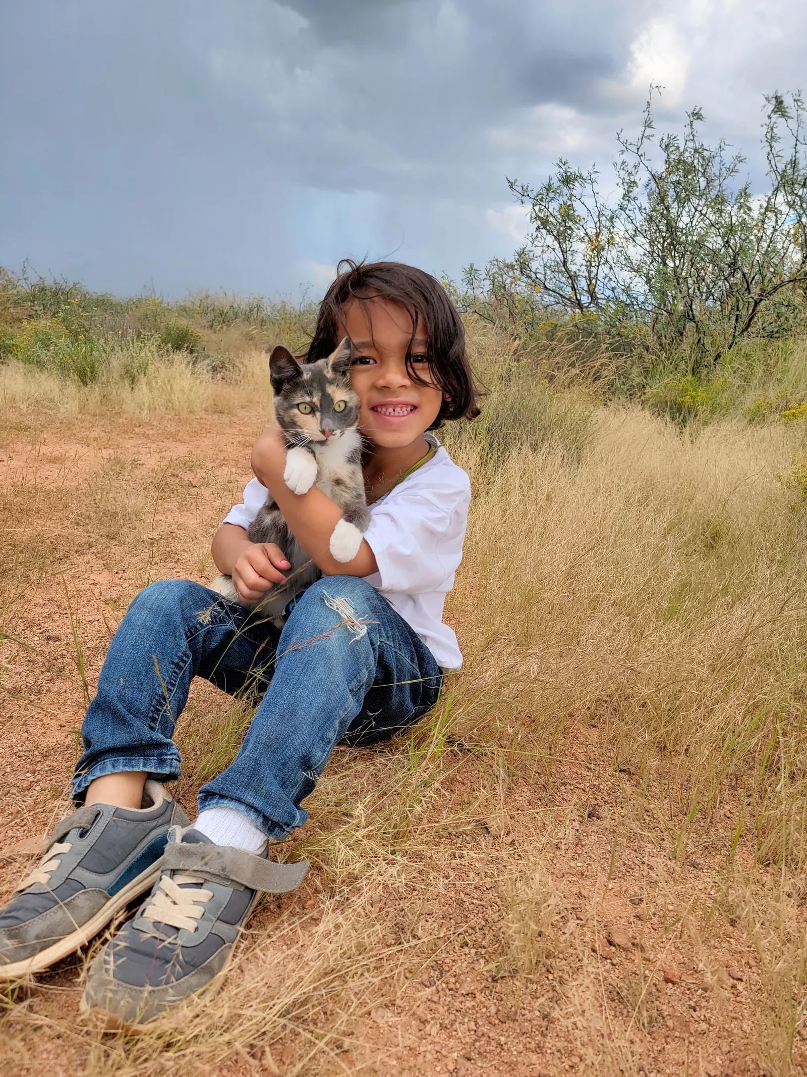 Child sitting in tall dry grass holding a cat on our homestead