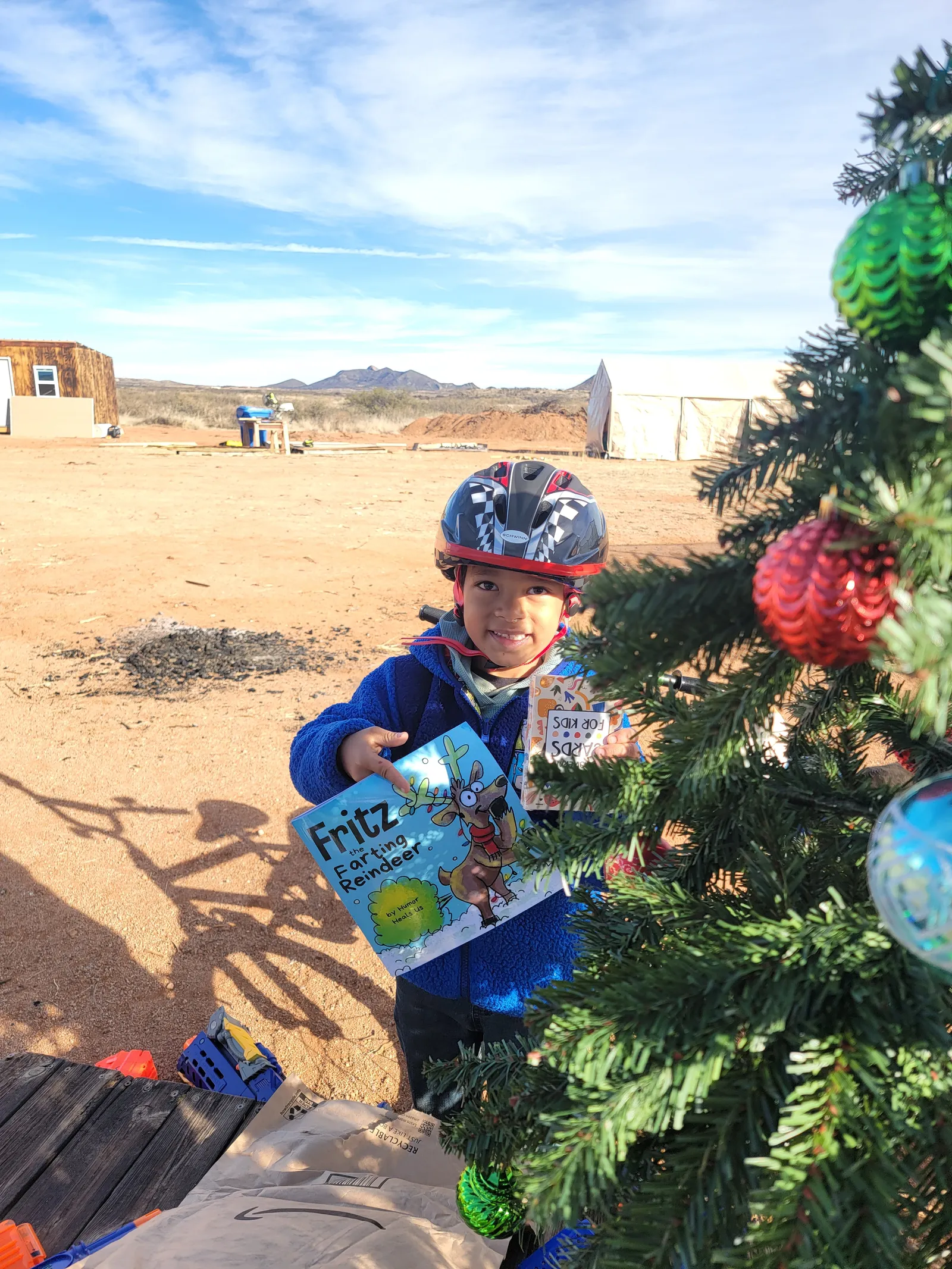 Child in a helmet holding a gift beside a decorated Christmas tree outdoors in Arizona