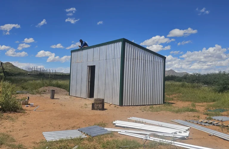 Metal shed with green trim on bare ground under a blue sky, a person working on the roof, on our off-grid homestead near Tombstone, Arizona