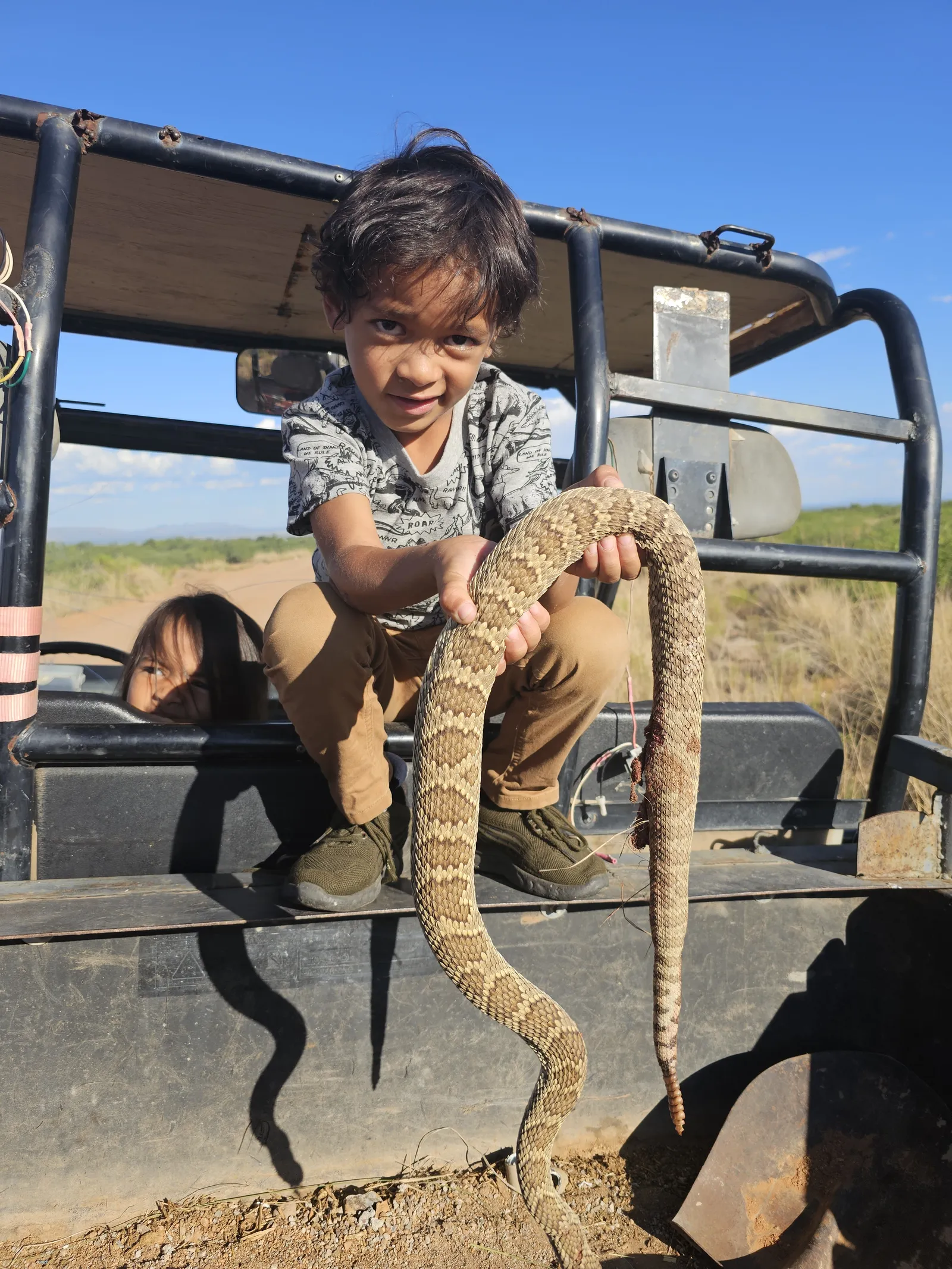 Young boy crouching on a utility vehicle holding a long snake, with another child looking out behind him