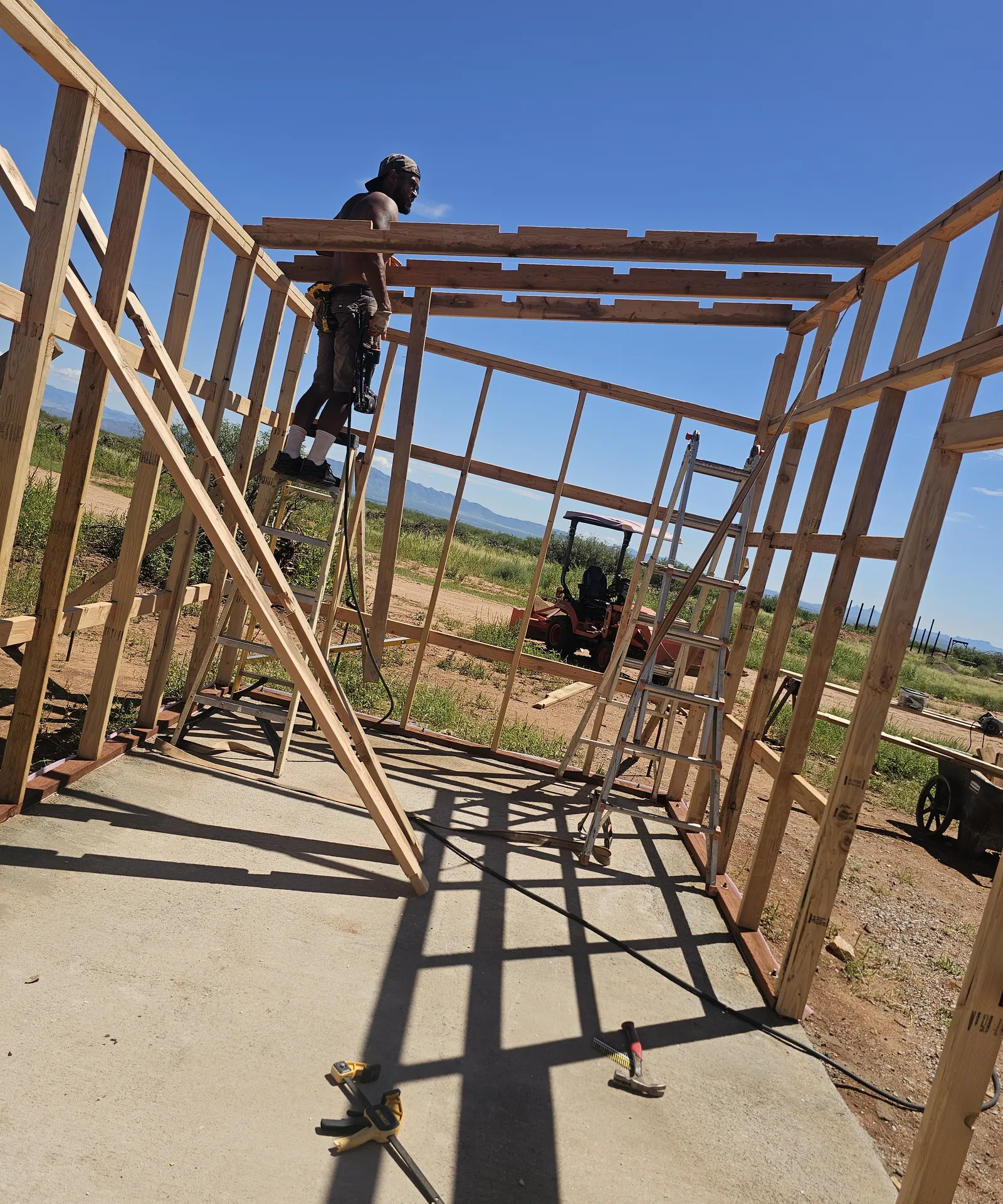 Wood shed framing with a person standing on top of the wall frame setting roof beams, ladder against the side